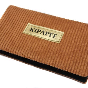 May include: A rectangular, corduroy wallet in a warm brown hue. The wallet features a black border and a gold label with the text "KIPAPEE". The corduroy texture adds a tactile element to the design.