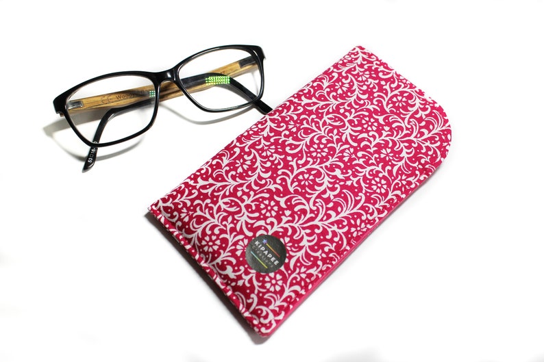 May include: A pink and white patterned glasses case with a white logo that says "Kipapee Designs".