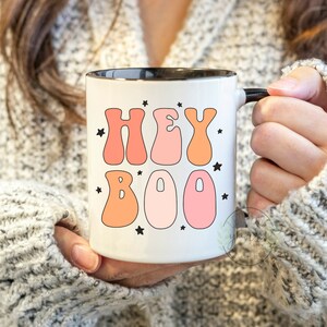 May include: White ceramic mug with a black rim and handle. The mug has a pink and orange graphic that says "Hey Boo" with black stars.