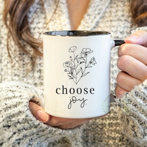 May include: White ceramic mug with a black rim and handle. The mug has a black line drawing of flowers and the text "choose joy."
