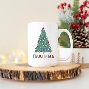May include: White ceramic mug with a green Christmas tree design and the text "Fa La La La La" in red and green. The mug is sitting on a wooden coaster.