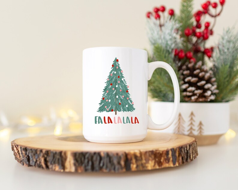 May include: White ceramic mug with a green Christmas tree design and the text "Fa La La La La" in red and green. The mug is sitting on a wooden coaster.