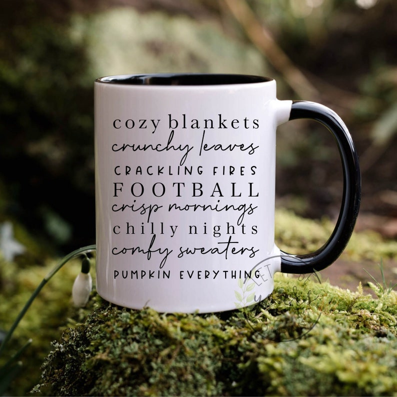 May include: White ceramic mug with a black handle and black rim. The mug has black text that reads "cozy blankets crunchy leaves crackling fires football crisp mornings chilly nights comfy sweaters pumpkin everything".