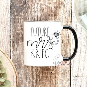 May include: White ceramic mug with a black handle. The mug has black text that reads "Future Mrs. Krieg" with a diamond ring illustration.