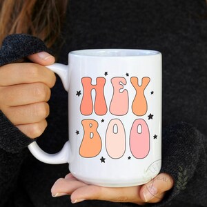 May include: White ceramic coffee mug with a pink and orange "Hey Boo" graphic and black stars.