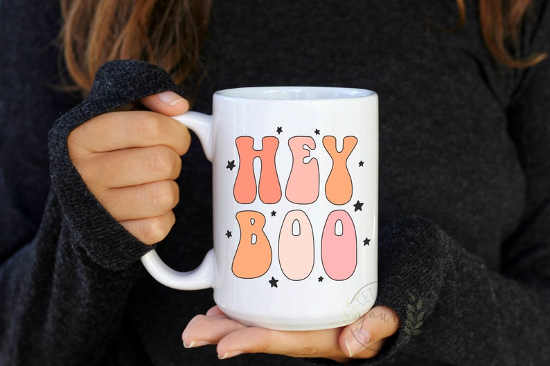 May include: White ceramic coffee mug with a pink and orange "Hey Boo" graphic and black stars.