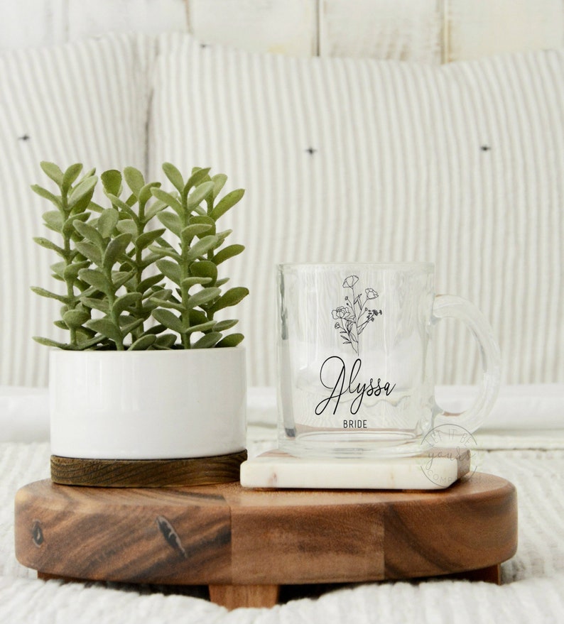 May include: A clear glass mug with a black floral design and the name "Alyssa Bride" printed on it. The mug is sitting on a white marble coaster on a wooden tray. A small potted succulent plant is on the left side of the tray.