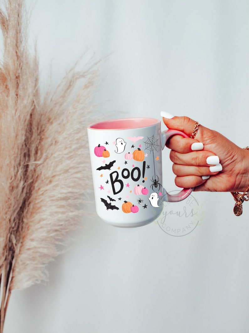 Peut inclure: Une tasse en c&eacute;ramique blanche avec un bord et une poign&eacute;e roses. La tasse pr&eacute;sente un motif d'Halloween avec des chauves-souris noires, des fant&ocirc;mes, des araign&eacute;es et des citrouilles. Le texte "Boo!" est &eacute;crit en noir avec des &eacute;toiles roses.