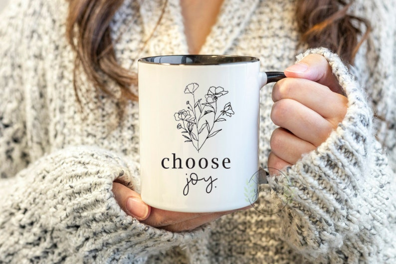 May include: White ceramic mug with a black rim and handle. The mug has a black line drawing of flowers and the text "choose joy."
