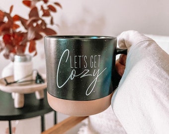 May include: Black ceramic mug with a brown rim and the text "Let's Get Cozy" in white lettering. The mug is being held by a hand wearing a white sweater.