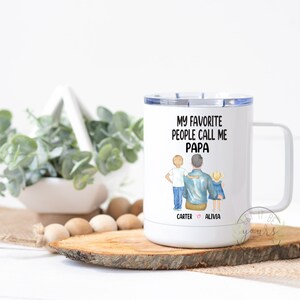 Op de afbeelding: Witte reistumbler van roestvrij staal met een zwart-blauwe illustratie van een vader en twee kinderen. De tekst "Mijn favoriete mensen noemen me Papa" staat op de beker gedrukt. De namen "Carter" en "Alivia" staan onder de illustratie gedrukt.