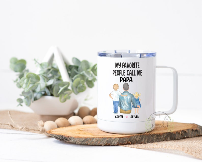Op de afbeelding: Witte reistumbler van roestvrij staal met een zwart-blauwe illustratie van een vader en twee kinderen. De tekst "Mijn favoriete mensen noemen me Papa" staat op de beker gedrukt. De namen "Carter" en "Alivia" staan onder de illustratie gedrukt.