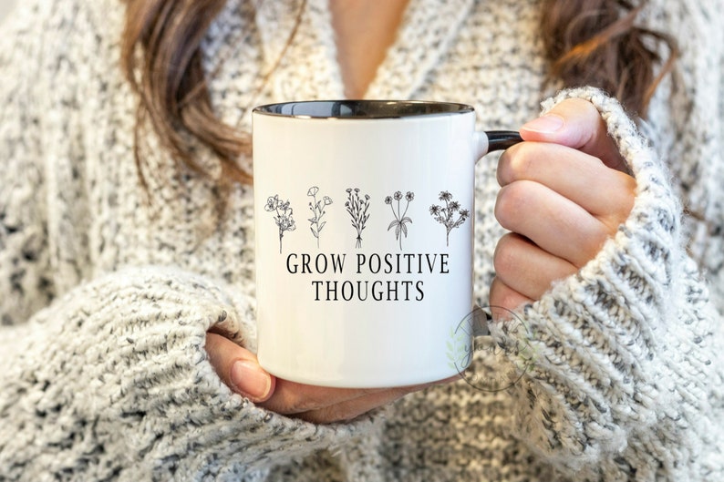 May include: White ceramic mug with a black rim and handle. The mug has a black line drawing of five flowers and the text "GROW POSITIVE THOUGHTS".