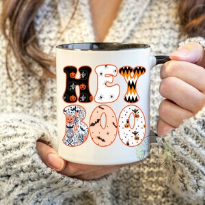 May include: A white ceramic mug with a black rim and handle. The mug has a Halloween-themed design with the words "Hey Boo" in colorful letters with various Halloween patterns.