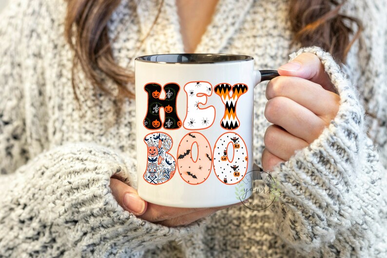 May include: A white ceramic mug with a black rim and handle. The mug has a Halloween-themed design with the words "Hey Boo" in colorful letters with various Halloween patterns.