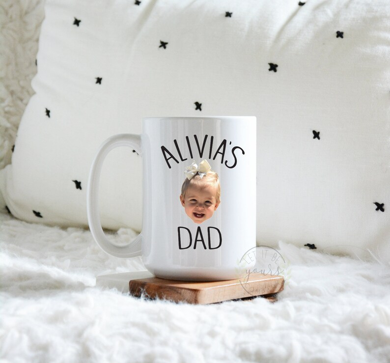 Peut inclure: Une tasse en c&eacute;ramique blanche avec une anse noire. La tasse est d&eacute;cor&eacute;e d'une photo d'un jeune enfant avec un n&oelig;ud sur la t&ecirc;te et le texte "Alivia's Dad" imprim&eacute; dessus.