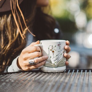 Pu&ograve; includere: Una tazza in ceramica bianca con un design floreale e la parola "remarkable" stampata su di essa. La tazza presenta un'illustrazione stilizzata di una persona che legge un libro.