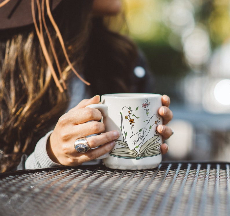 Pu&ograve; includere: Una tazza in ceramica bianca con un design floreale e la parola "remarkable" stampata su di essa. La tazza presenta un'illustrazione stilizzata di una persona che legge un libro.