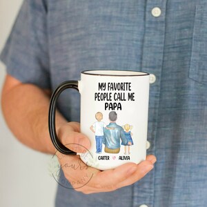 Op de afbeelding: Een witte keramische mok met een zwart handvat. De mok heeft een zwarte rand en een ontwerp dat zegt "Mijn favoriete mensen noemen me Papa" met een cartoonillustratie van een vader en twee kinderen. De namen van de kinderen zijn "Carter" en "Alivia" met een hartje ertussen.