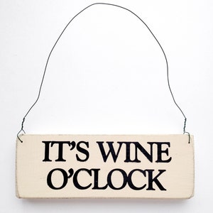 May include: A white wooden sign with black lettering that reads "It's Wine O'Clock". The sign is hanging from a wire.