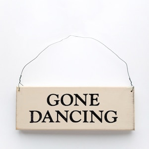 May include: A white wooden sign with black text that reads "Gone Dancing" hanging from a wire.