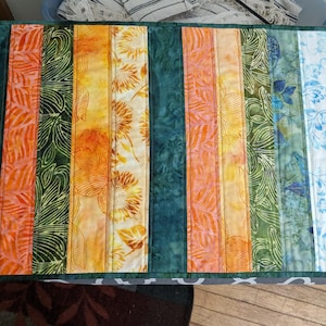 May include: A colorful table runner with vertical stripes of various patterns and colors. The stripes include orange, yellow, green, and blue floral and leaf designs. The edges are a dark green.