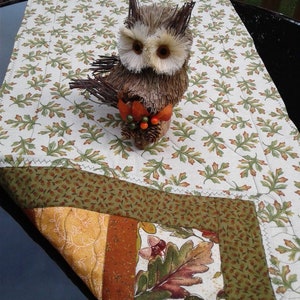 May include: A brown and white owl figurine made of twigs and straw, with a small cluster of red and orange berries. The owl is sitting on a white tablecloth with a green and brown leaf pattern. The tablecloth is folded over a dark blue surface.