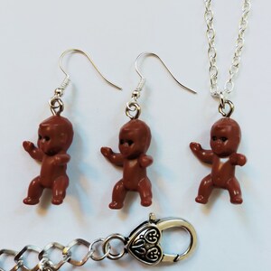 May include: A set of brown baby doll earrings and a necklace. The earrings and necklace are silver-toned and feature a brown baby doll charm.