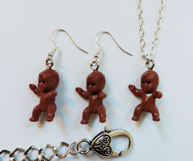 May include: A set of brown baby doll earrings and a necklace. The earrings and necklace are silver-toned and feature a brown baby doll charm.