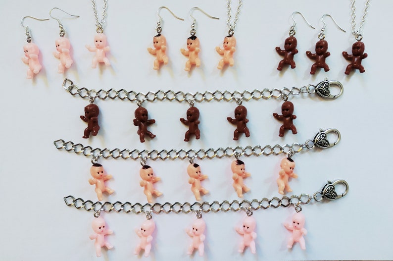 May include: Three silver chain bracelets with baby doll charms. The bracelets have heart-shaped clasps. The charms are pink, brown, and nude.