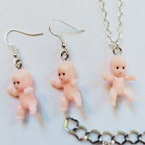 May include: A set of pink baby doll earrings and a necklace. The earrings and necklace feature a small plastic baby doll charm.