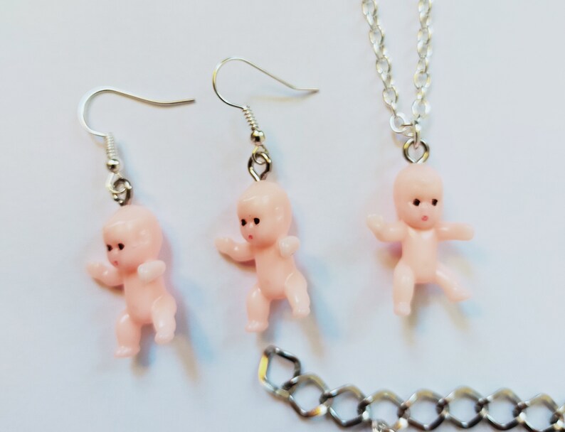 May include: A set of pink baby doll earrings and a necklace. The earrings and necklace feature a small plastic baby doll charm.