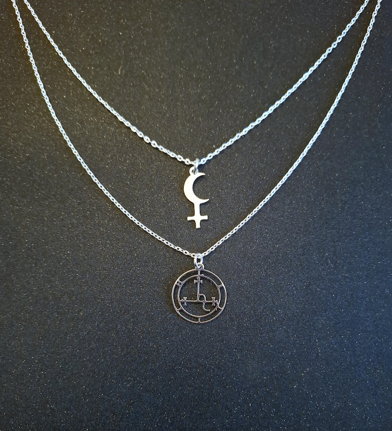 May include: Two silver necklaces with pendants. The top necklace has a crescent moon and a cross pendant. The bottom necklace has a circular pendant with a symbol inside.
