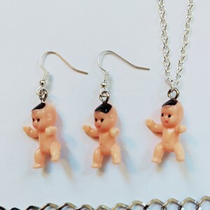 May include: A set of silver baby doll earrings and a necklace. The dolls are pink with black hair and are wearing silver earrings.