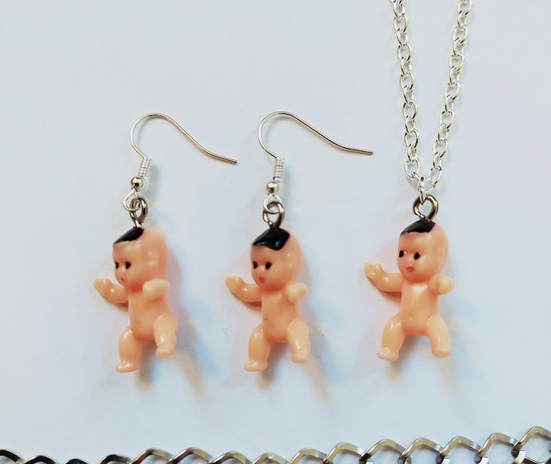May include: A set of silver baby doll earrings and a necklace. The dolls are pink with black hair and are wearing silver earrings.