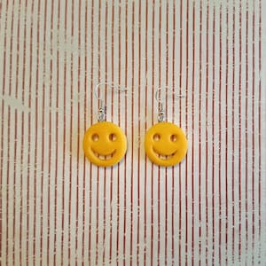 Peut inclure: Deux boucles d'oreilles smiley jaunes. Chaque boucle d'oreille présente un visage rond et souriant avec des détails des yeux et de la bouche. Les boucles d'oreilles sont suspendues à des crochets d'oreille argentés. Le fond est un motif à rayures blanches et marron.