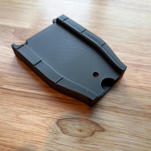 Op de afbeelding: Een donkergrijs, gebogen kunststof onderdeel met een getextureerd oppervlak en een klein gaatje. Het object heeft een rechthoekige vorm met afgeronde randen, tegen een houten achtergrond.