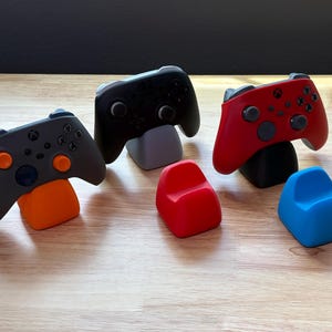 May include: Three video game controllers resting on individual stands, including a gray controller on an orange stand, a black controller on a gray stand, and a red controller on a black stand, plus two empty stands in red and blue.