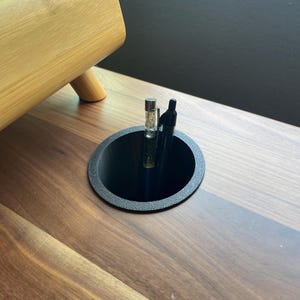 May include: A black plastic pen holder with a round opening, sitting on a wooden desk. Three pens are inside the holder, including a clear pen with glitter inside.