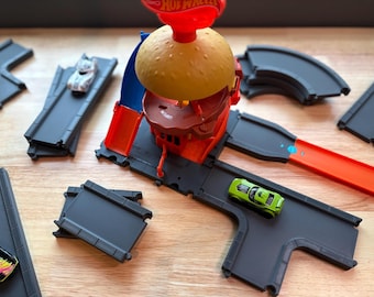 May include: A Hot Wheels burger-themed playset featuring a yellow bun top, a red base, and multiple gray plastic track segments. A small white toy car sits on one track, while a bright green toy car is positioned on another track section.