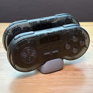 Peut inclure: Deux manettes de jeu 8BitDo gris foncé translucides empilées sur un petit support gris clair. Chaque manette comprend une croix directionnelle, quatre boutons d'action circulaires, deux boutons centraux Select et Start, et des boutons de tranche.