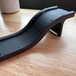 May include: A black plastic toy bridge with a curved design. The bridge has a flat surface for toy cars or trains to cross. The bridge is elevated by a single support. The toy bridge is on a wooden surface.