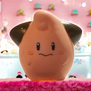 May include: A light peach-colored, cartoon-style figure with black ear-like features and small, black eyes. The figure has a small, curved mouth and pink blush marks on its cheeks. It is sitting on a pink, fuzzy surface.