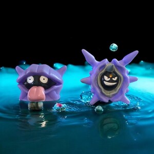 May include: Two purple Pokémon figures, one resembling a clam with a pink tongue and the other a spiky, round shape with a smiling face. The figures are set against a blue water background with a black backdrop.