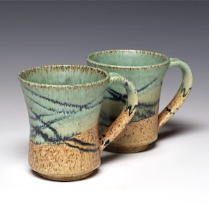 May include: Two handmade ceramic mugs. The mugs have a light green and tan color scheme with black accent lines. The handles are curved and the rims are slightly flared. The mugs are likely for coffee or tea.
