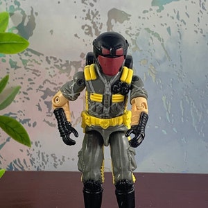 May include: A gray and black action figure with a yellow belt and a red face mask. The figure is wearing a black helmet and has a backpack.
