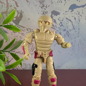 May include: A beige action figure with a helmet and a red and black belt. The figure is wearing a beige and black suit of armor. The figure is standing on a dark red surface.