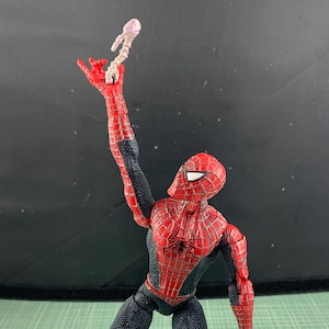 May include: A Spider-Man action figure in a dynamic pose, clad in a red and black suit. The figure is holding a small, light-colored object in its raised hand. The figure's pose suggests action, with its legs spread and arms extended.