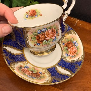May include: A vintage teacup and saucer set featuring a floral design. The cup and saucer are white with a blue and gold trim. The floral design includes pink and red roses with green leaves.
