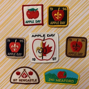 Peut inclure: Plusieurs écussons brodés sur le thème de la pomme. Les écussons présentent des pommes rouges, le texte "APPLE DAY" et les années 1994, 1995, 1996 et 1997. Certains écussons incluent également une fleur de lys ou une feuille d'érable.